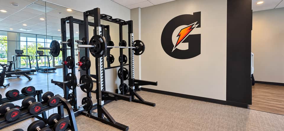 Weight training area with squat racks, barbell plates, and Gatorade branded wall in bright gym space