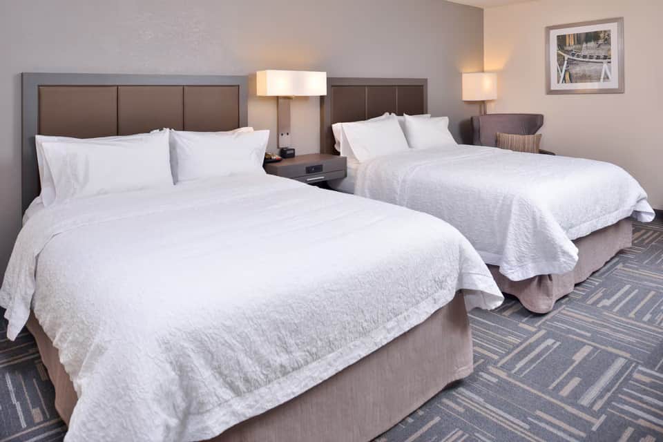 Two queen beds with white bedding in a modern hotel room with brown headboards and patterned carpet