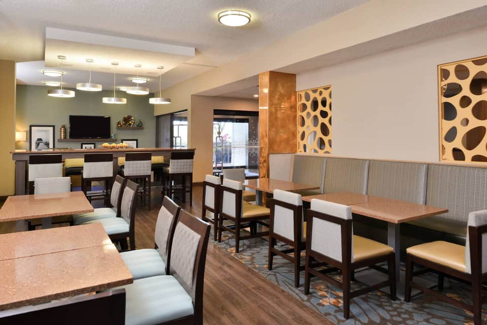 Hotel breakfast area with dining tables, chairs, bar seating, and decorative wall art with ambient lighting