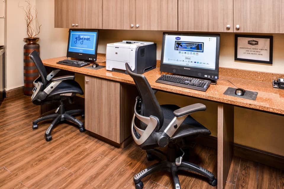 Business center with two ergonomic chairs, dual monitors, printer, and wooden desk in hotel office