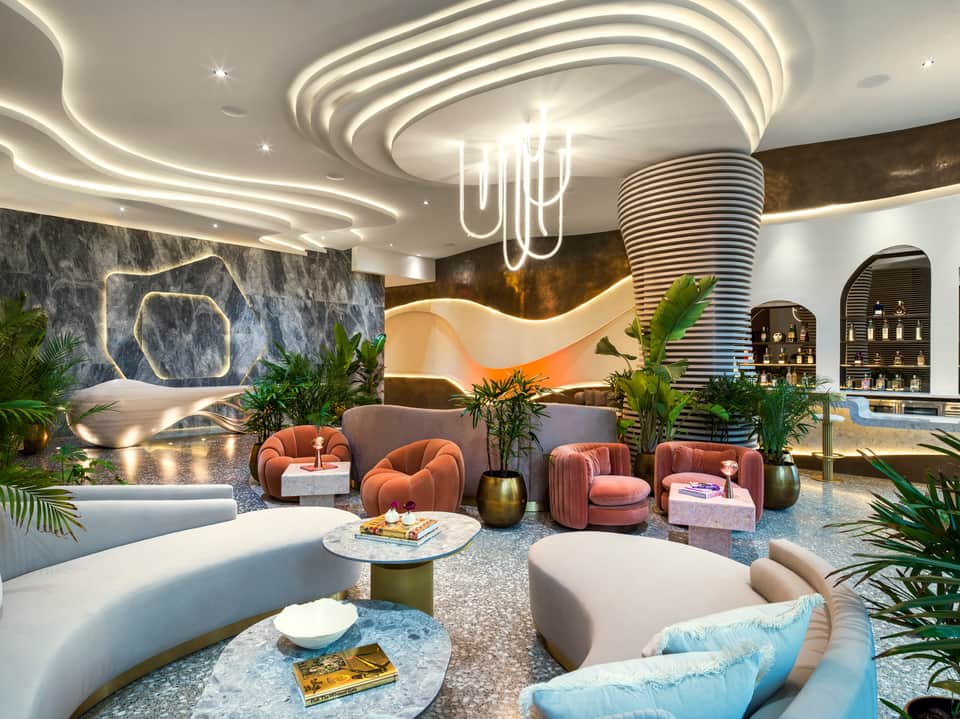Modern hotel lobby lounge with curved furniture, neon lighting, marble floors, and tropical plants
