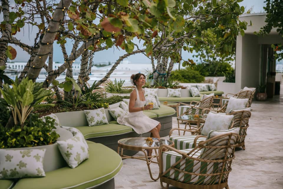 Woman relaxing on curved green lounge under twisted trees on oceanview terrace with rattan seating