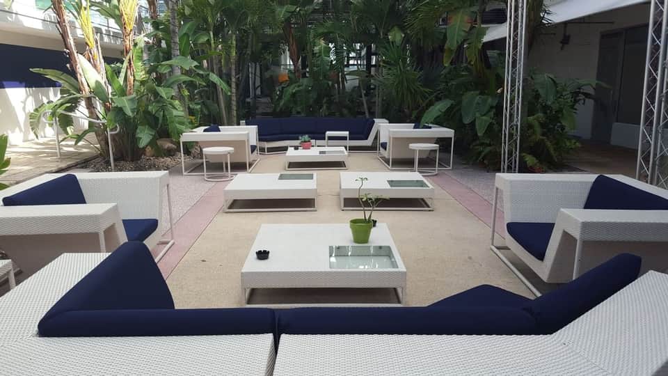 Modern outdoor lounge area with white wicker furniture, dark blue cushions, and lush tropical plants surrounding seating sections