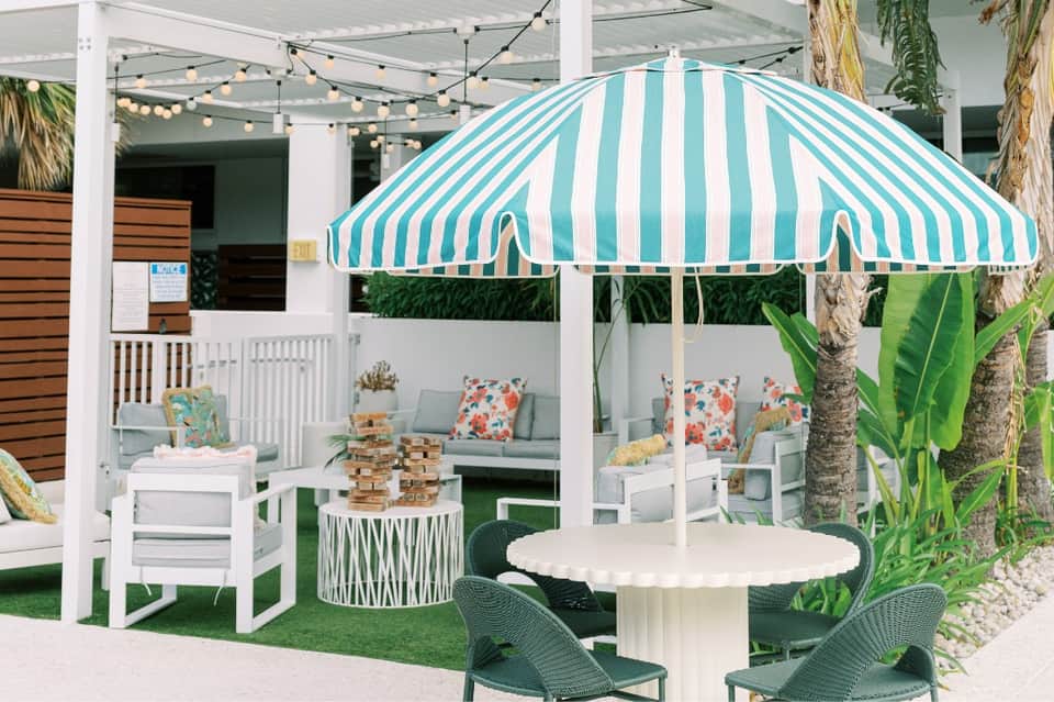 Tropical resort patio with turquoise striped umbrella, white furniture, and palm trees under string lights