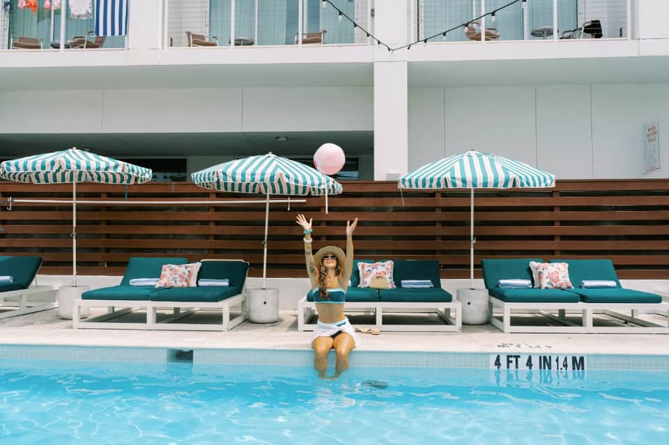 Woman sitting poolside under teal striped umbrellas with lounge chairs and modern hotel building