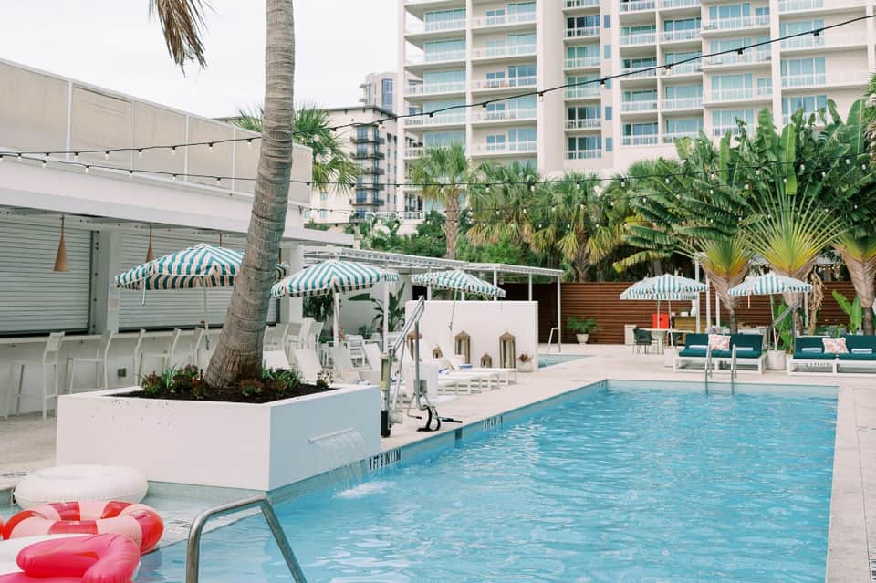 Resort pool surrounded by green-striped umbrellas, loungers, and multi-story hotel building