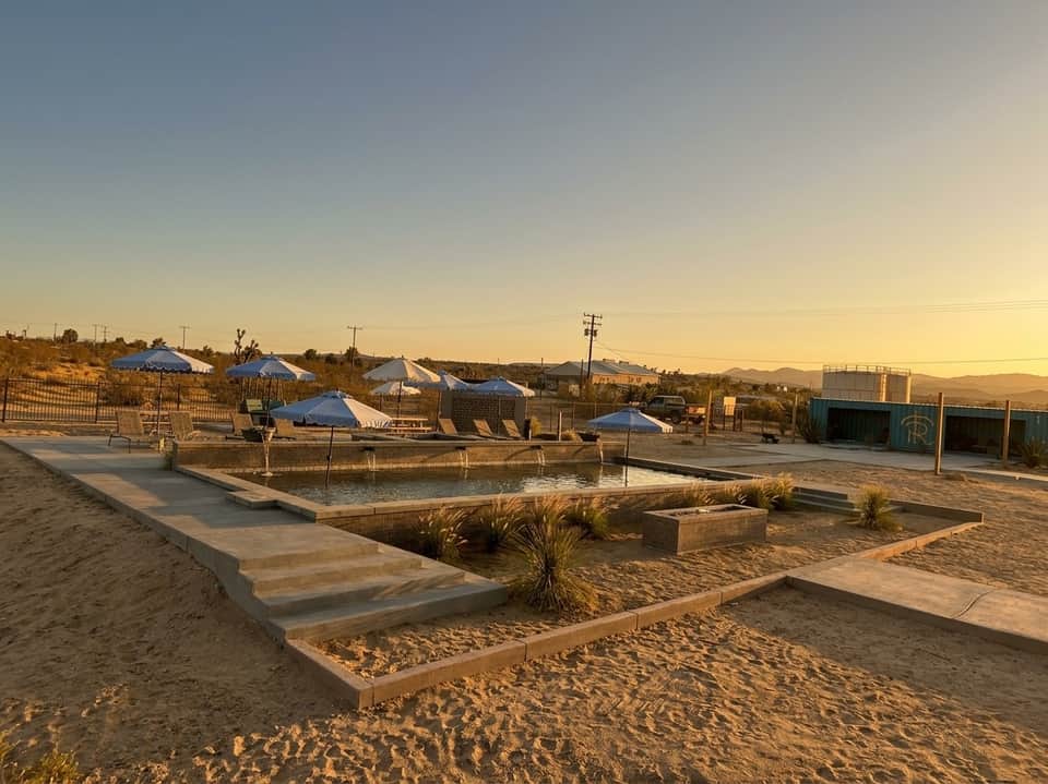 Desert resort at sunset with concrete pool deck, blue umbrellas, and sandy landscape with distant mountains