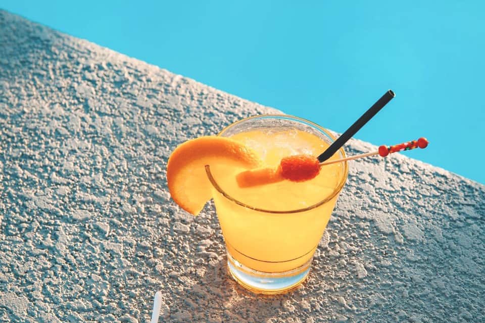 Yellow tropical cocktail with orange slice and cherry garnish on textured surface against blue sky