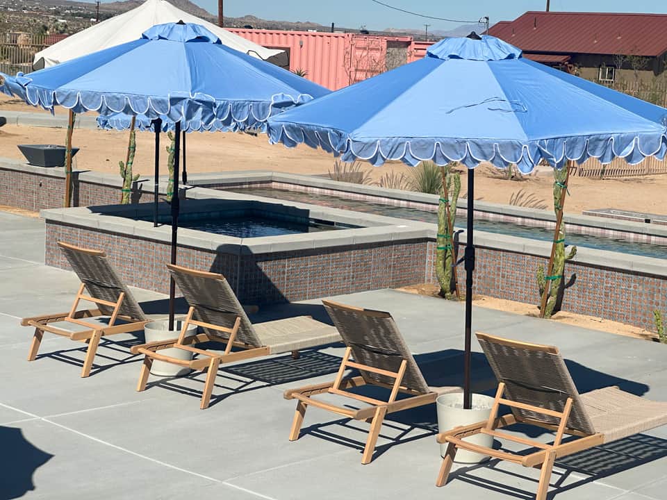 Two blue umbrellas over lounge chairs next to an empty pool basin in desert setting