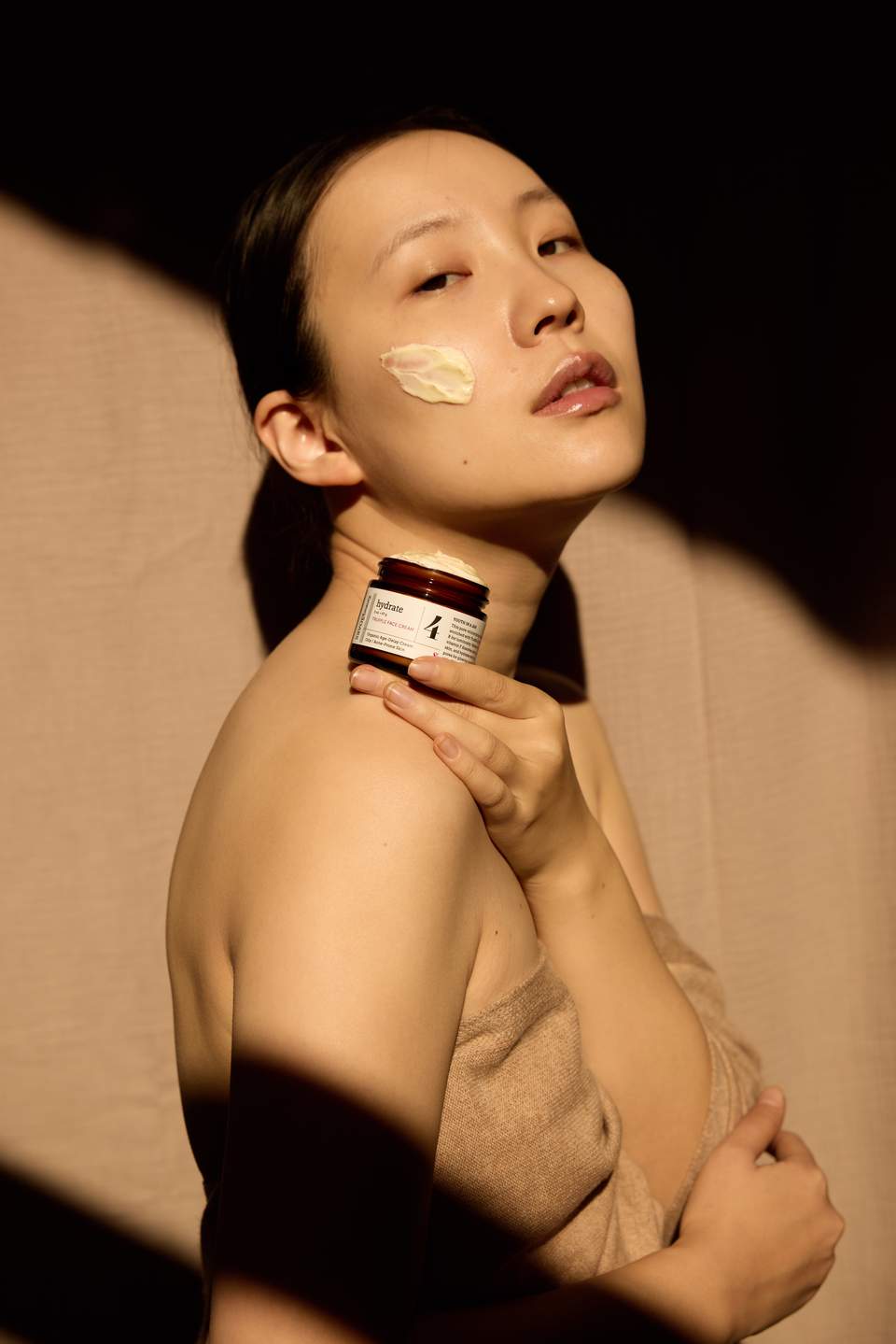 Woman applying cream to cheek while holding skincare jar with neutral background
