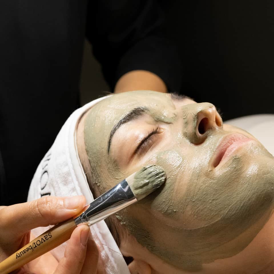 Esthetician applying green clay mask to client's face with cosmetic brush