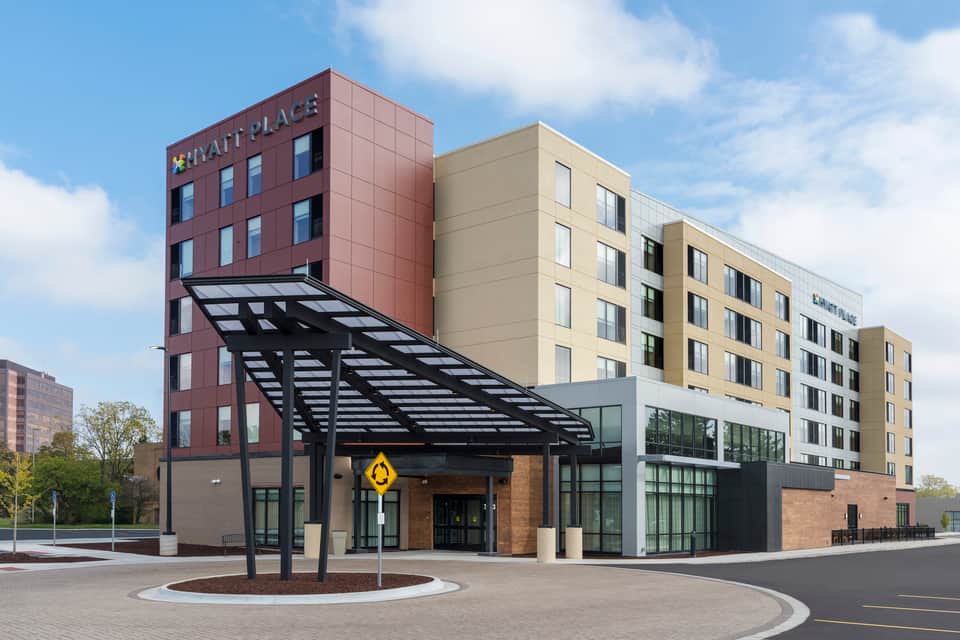 Modern Hyatt Place hotel exterior with burgundy tower, beige wings, black pergola entrance canopy, and circular courtyard
