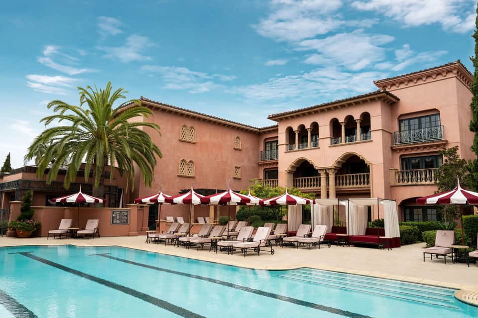 Mediterranean-style resort with pink stone building, pool, red and white umbrellas, lounge chairs, and palm trees