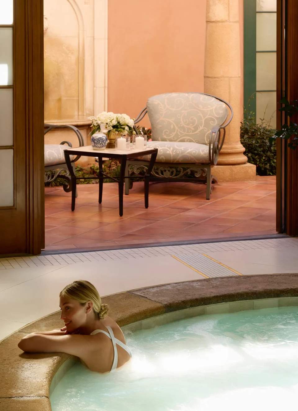 Woman relaxing in hot spring spa pool with courtyard seating area and decorative flowers in background