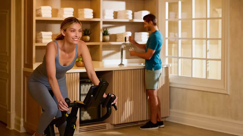 Woman on stationary bike in home gym with man getting water, wooden shelving with towels, modern aesthetic