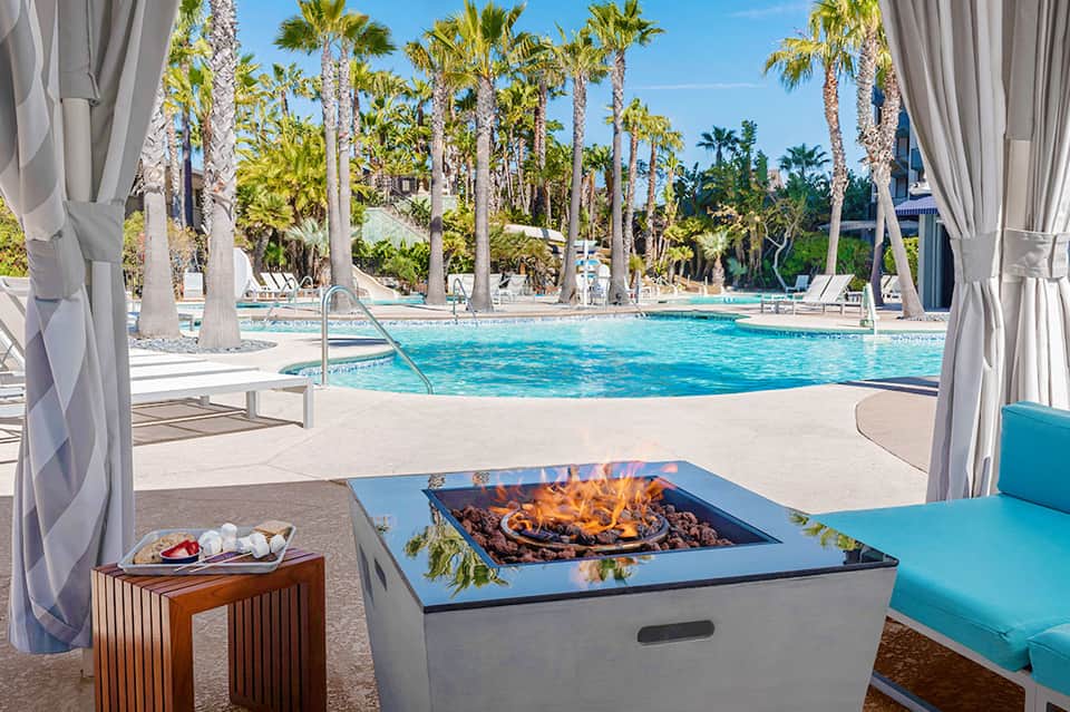 Luxury resort pool with fire pit, white cabanas, and tall palm trees under clear blue sky