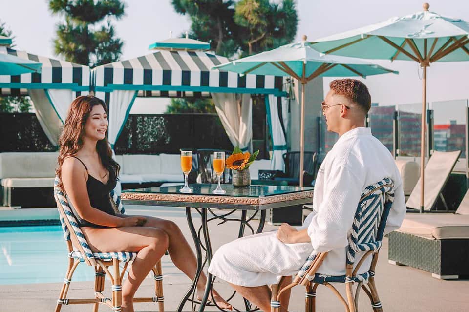 Couple enjoying drinks at a poolside table under turquoise umbrellas and striped cabanas.