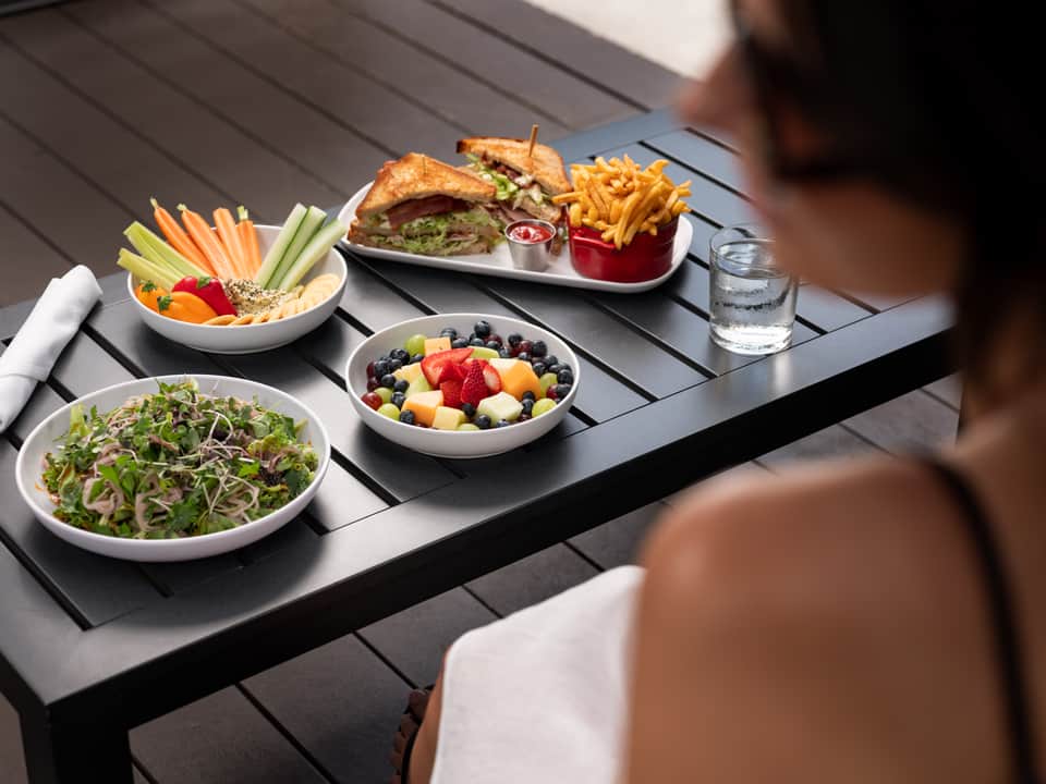 Outdoor dining table with fresh salads, burger, fruit bowl, and vegetable crudités on dark decking