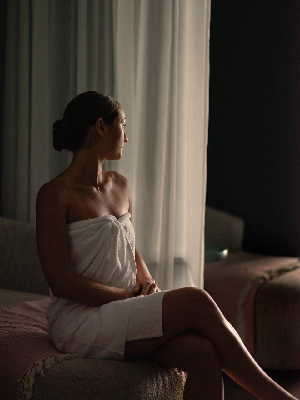 Woman in white towel sitting on bed by window with sheer curtains overlooking night sky