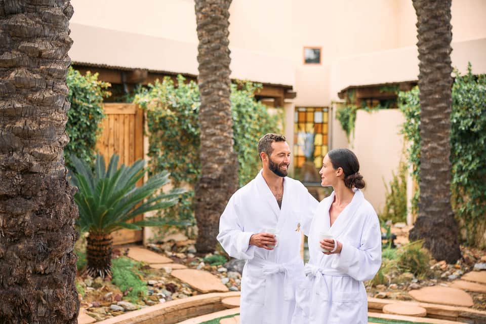 Couple in white robes walking through courtyard with palm trees, ivy, and warm architectural lighting