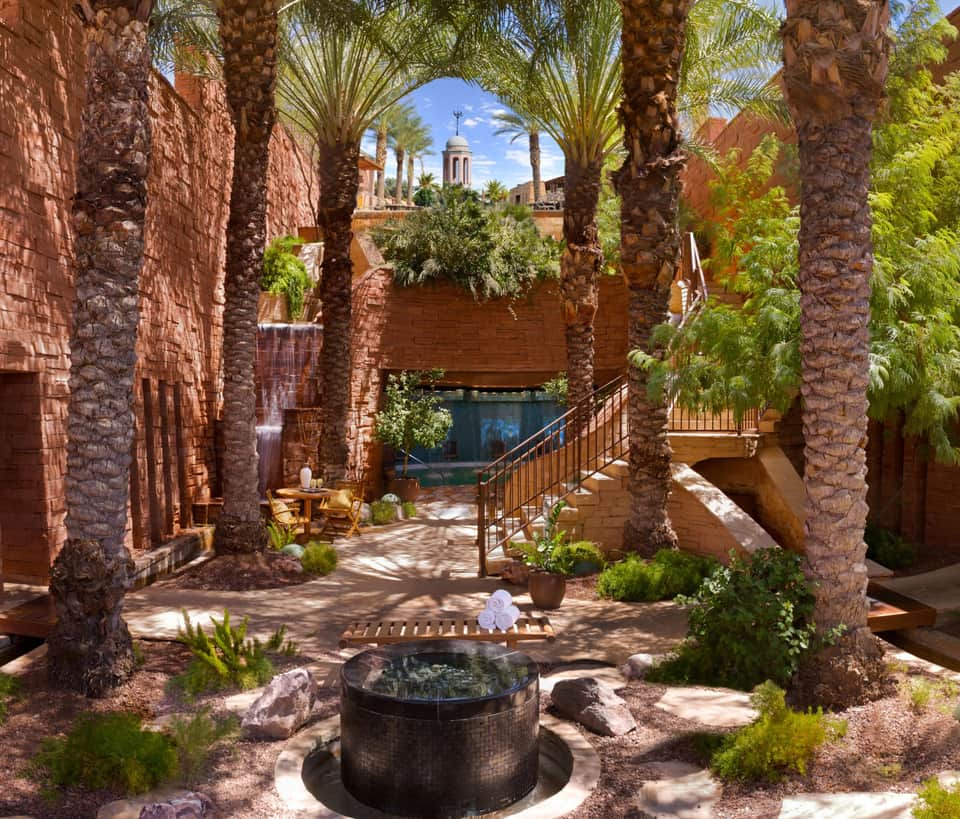 Spa courtyard with hot spring pool surrounded by red stone architecture, palm trees, and mountain views