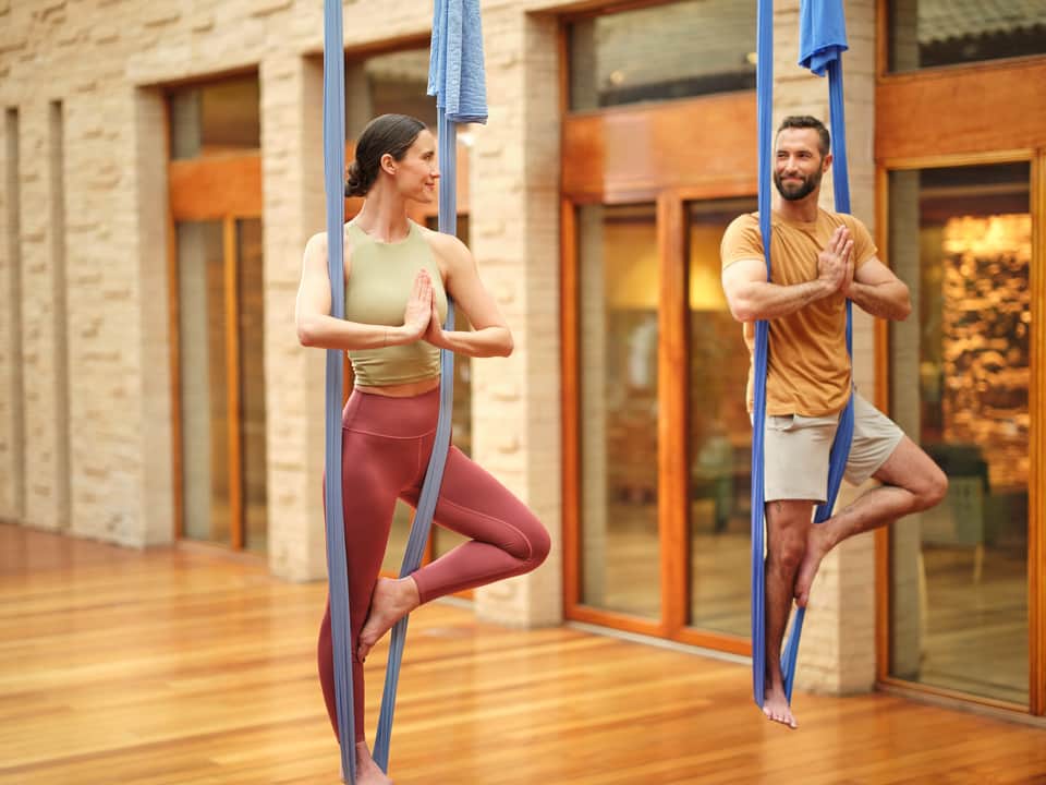 Man and woman performing aerial yoga poses using blue silk hammocks suspended in a modern courtyard with stone columns