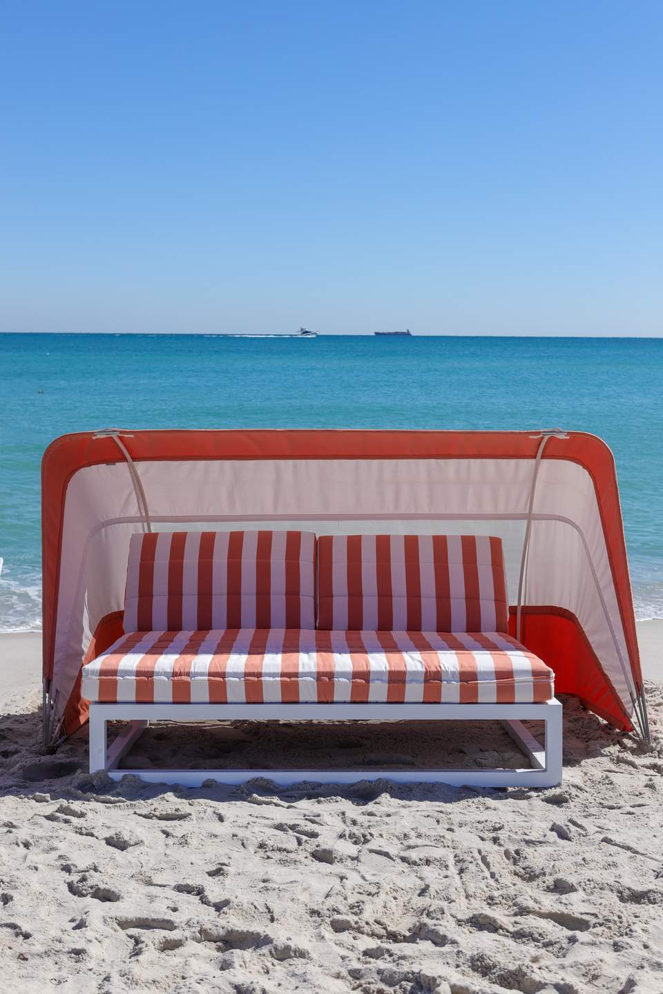 Red and white striped beach tent with cushioned seating on sand with ocean view