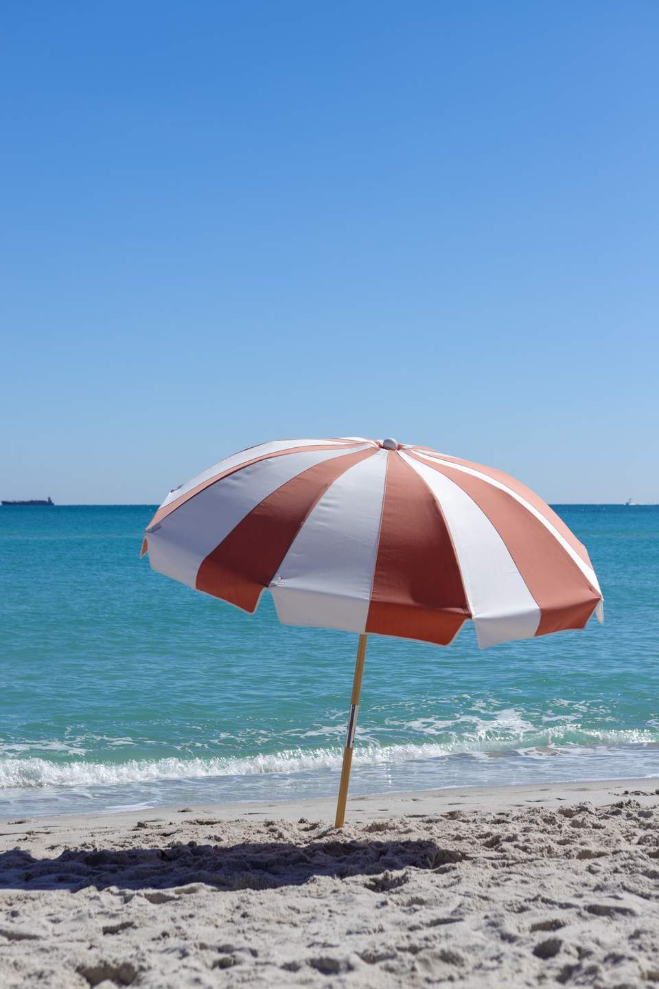 Red and white striped beach umbrella planted in sand overlooking ocean waves