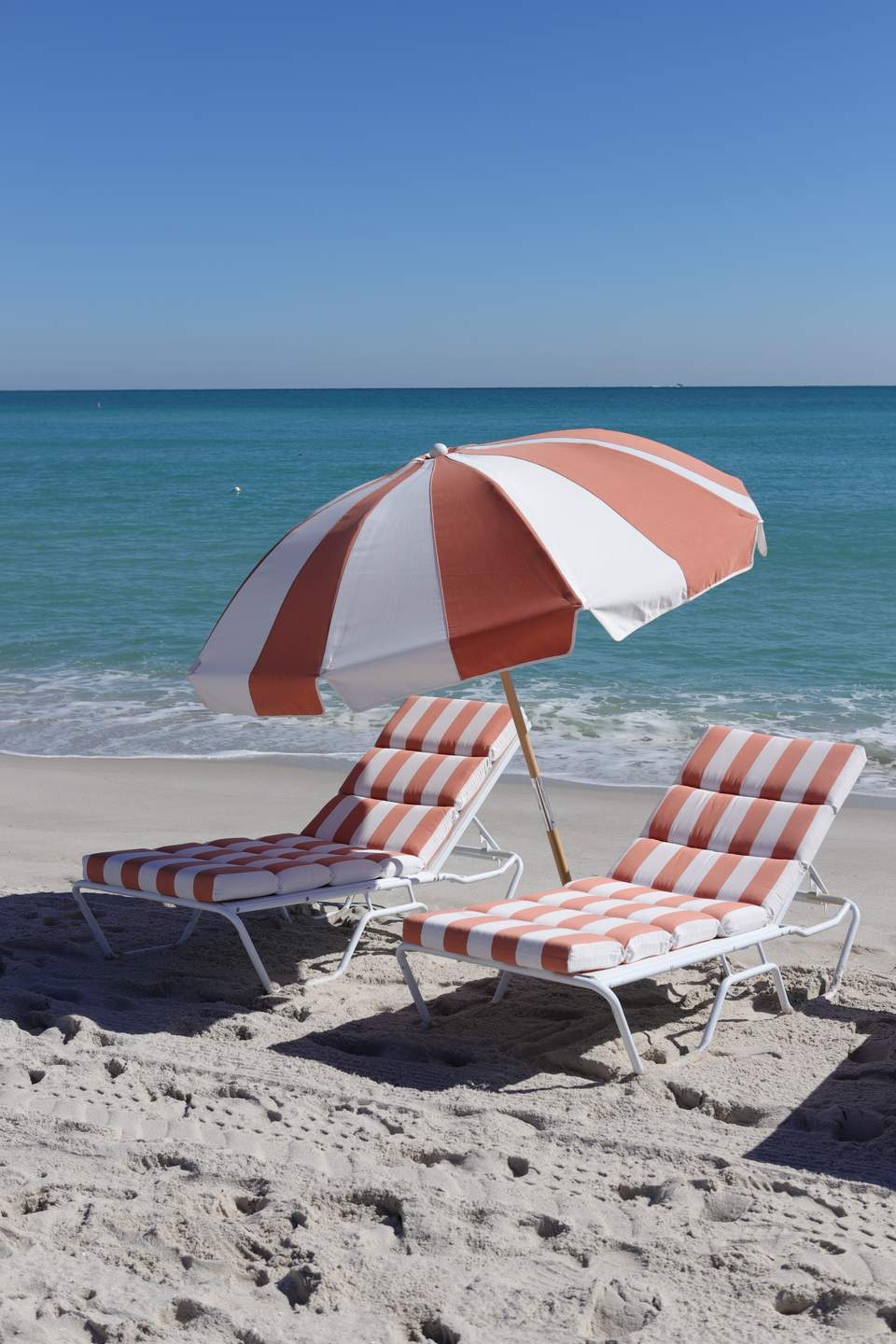 Two red and white striped beach loungers with umbrella on sandy beach facing ocean