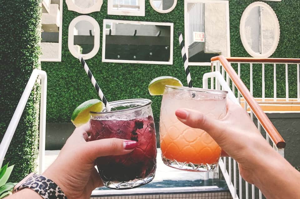 Two colorful cocktails with lime garnish held on a balcony overlooking green landscaping