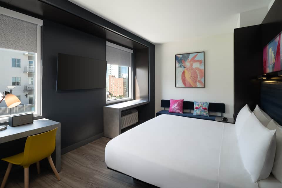 Hotel bedroom with queen bed, black accent wall, yellow desk chair, and city skyline views