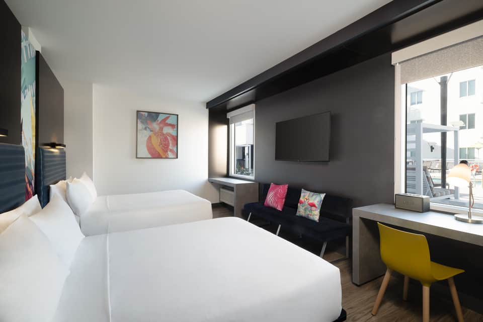 Twin beds in modern hotel room with black headboard, colorful pillows, desk area, and balcony access
