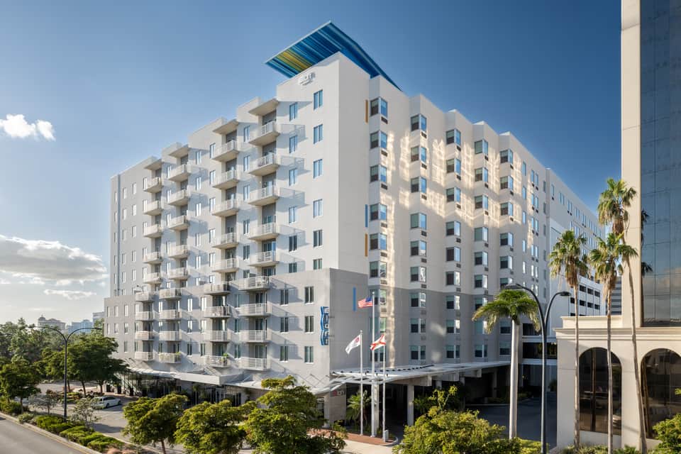 Eight-story white residential building with balconies, colorful roof accent, palm trees, and ground-level retail spaces