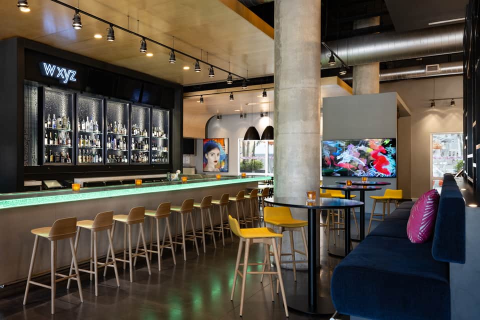 Hotel bar and lounge with illuminated bar counter, yellow chairs, TV screens, and contemporary design elements