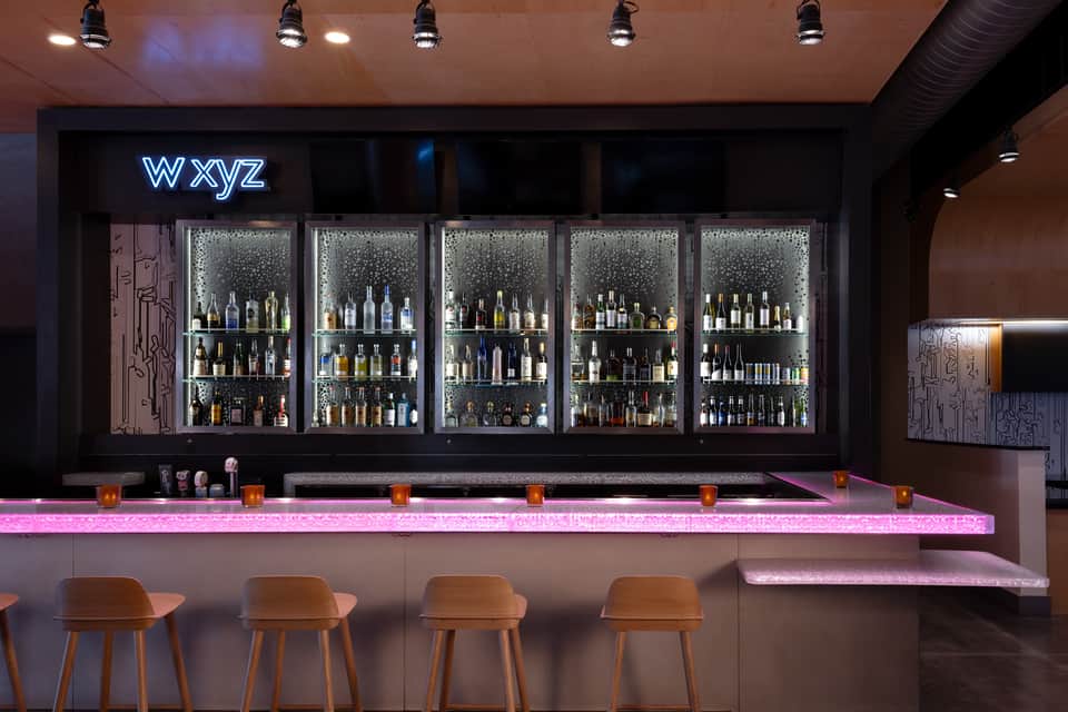 Modern bar interior with neon 'wxyz' sign, illuminated pink counter, backlit bottle display, and wooden bar stools