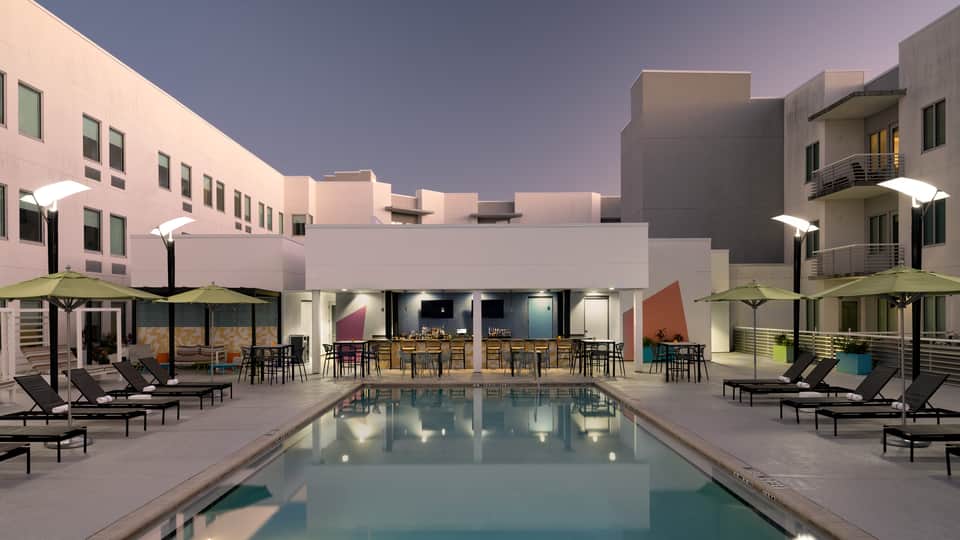 Resort courtyard at dusk with rectangular pool, lounge chairs, umbrellas, and contemporary multi-story buildings