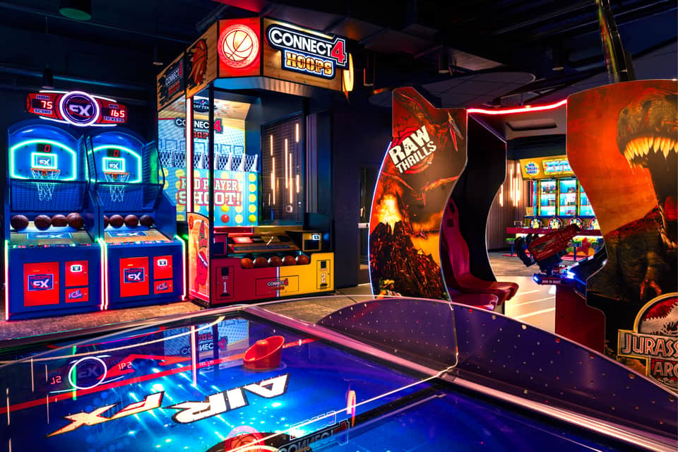 Arcade entertainment area featuring neon-lit basketball games, Connect Four, pinball, and dinosaur-themed games
