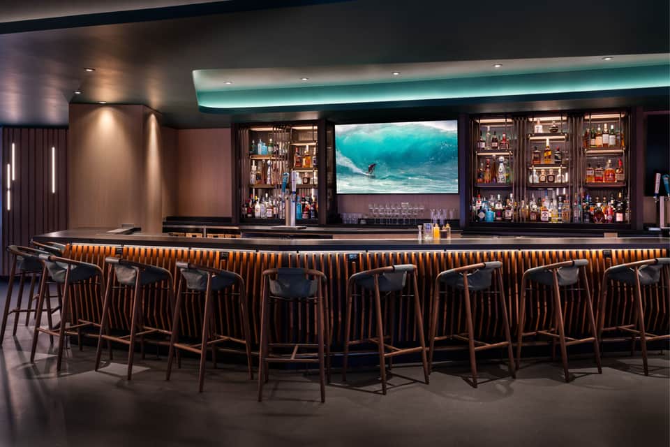 Modern hotel bar with wood-paneled seating, multiple TV screens displaying surfing content, and backlit shelving