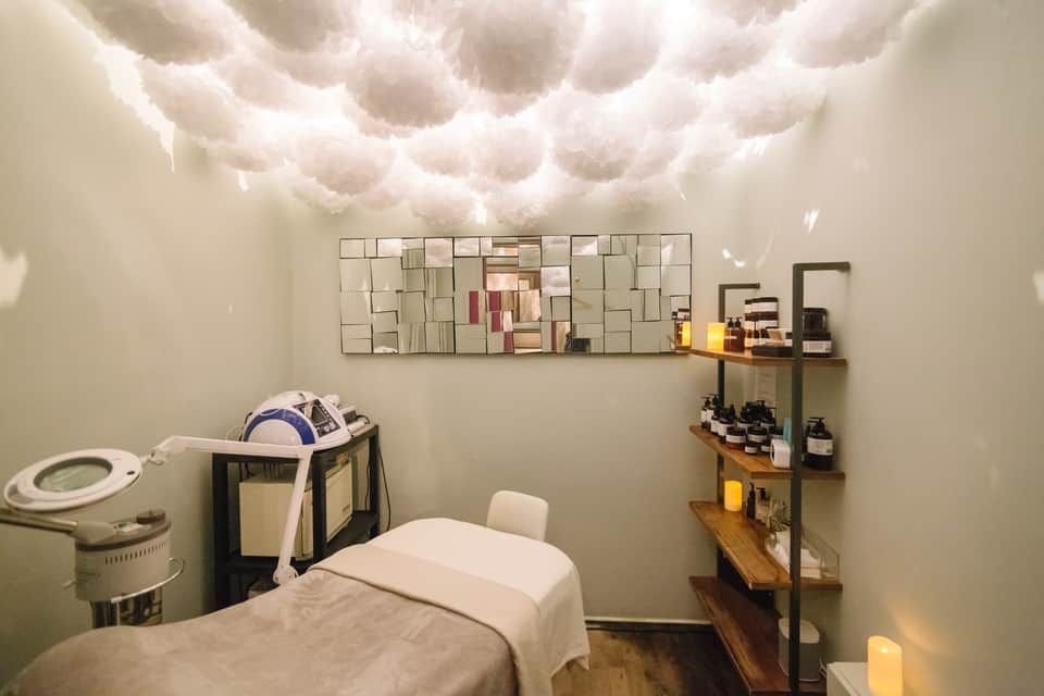 Spa treatment room with white puffy cloud ceiling installation, massage bed, and shelving with wellness products