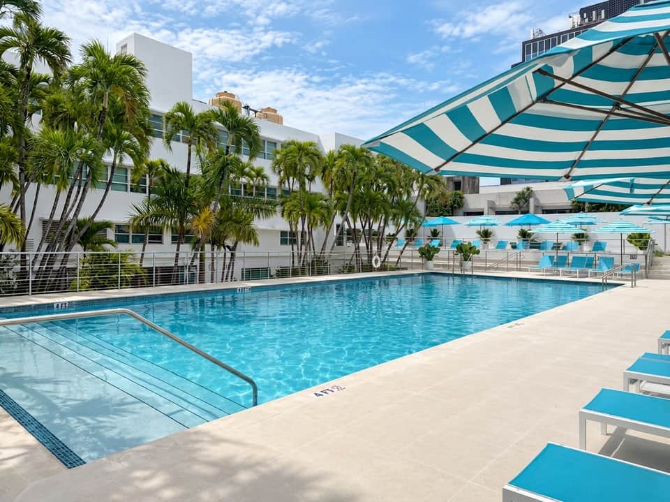 Sunny outdoor swimming pool with turquoise umbrellas, lounge chairs, palm trees, and white modern hotel building