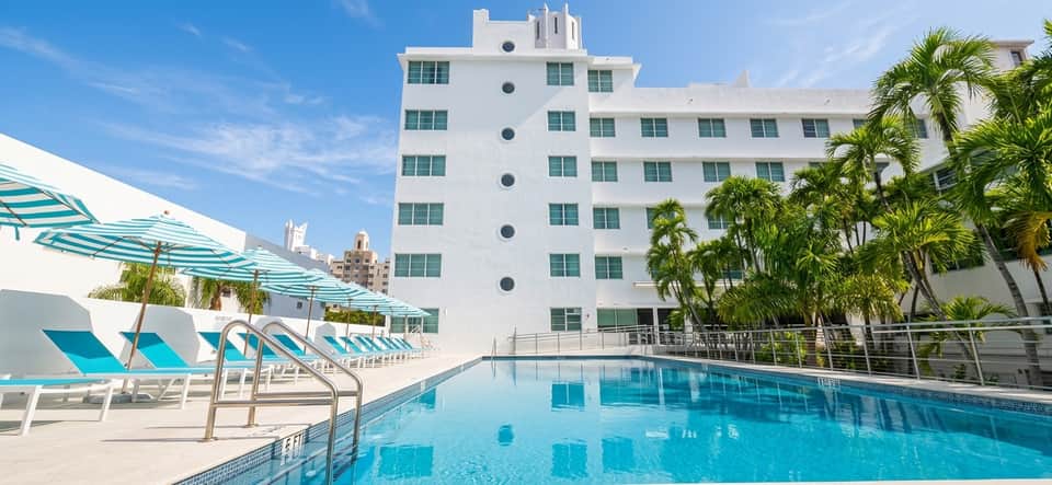 Resort pool with bright turquoise water, white loungers, umbrellas, palm trees, and Art Deco-style hotel tower