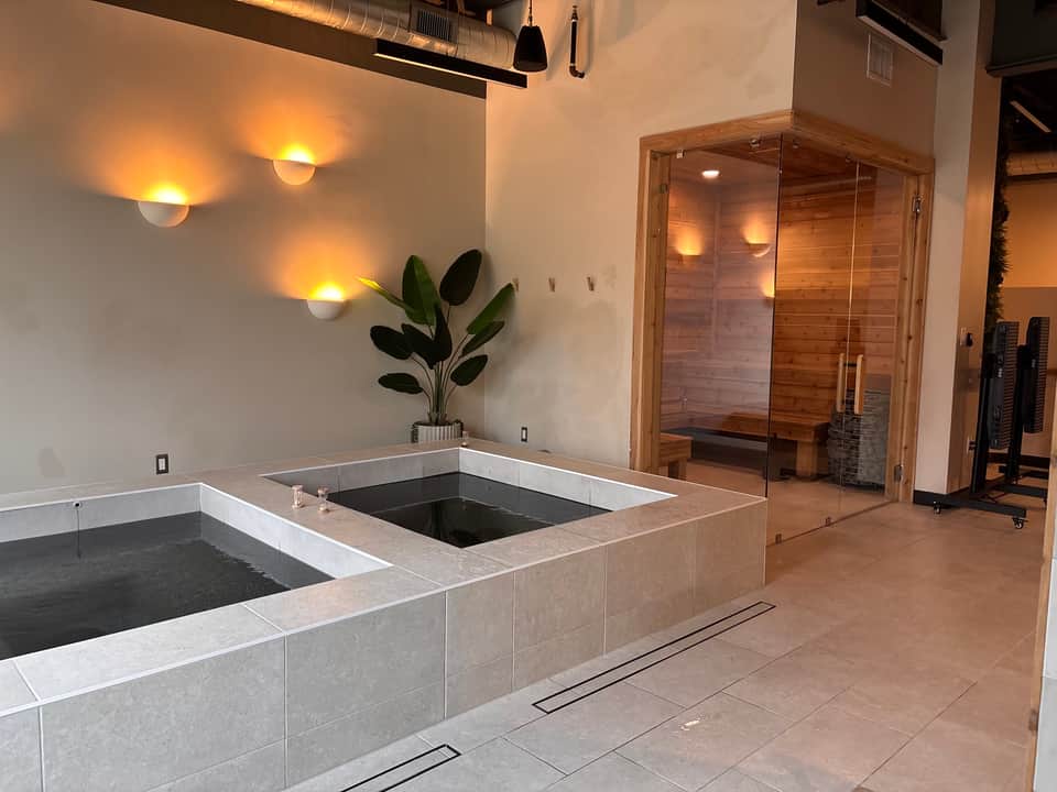 Spa relaxation area with two soaking tubs, wooden sauna with glass doors, warm accent lighting, and potted plant