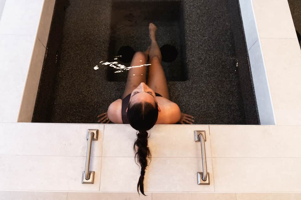 Overhead view of person floating in dark mineral water bath with arms raised