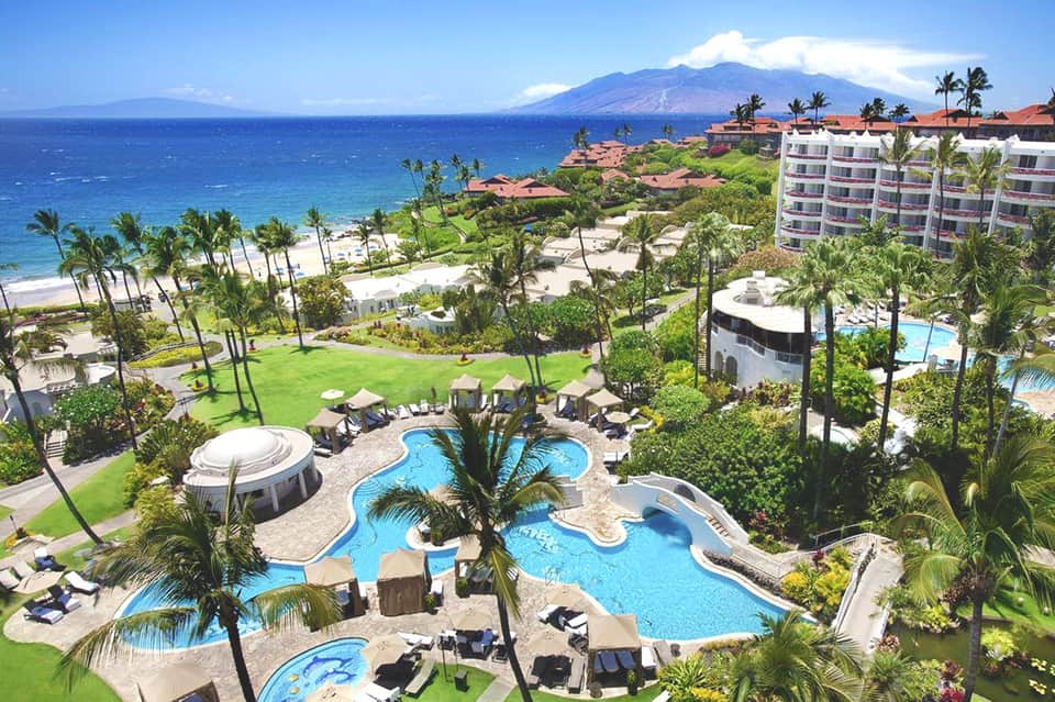 Aerial view of beachfront resort with curved pools, palm trees, and West Maui mountain in background