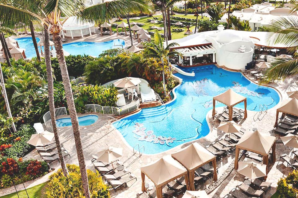 Aerial view of resort with curved blue pools, palm trees, lounge chairs, and umbrellas surrounded by manicured grounds