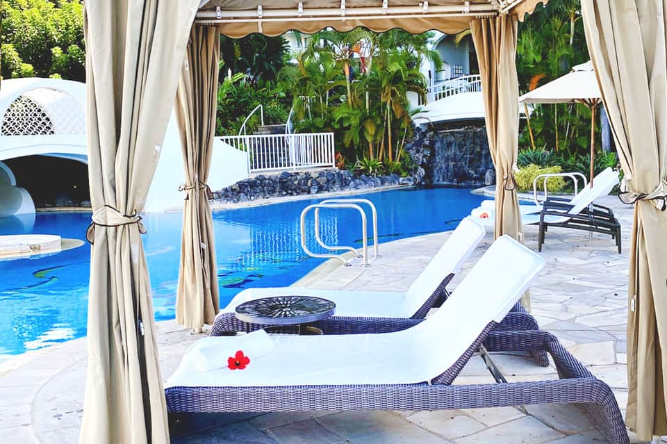 Pool cabana with white lounge chairs, cream curtains, and blue swimming pool