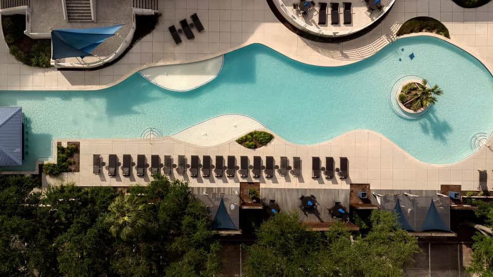 Aerial view of a luxury resort's curved turquoise pool with lounge chairs, umbrellas, and palm tree landscaping