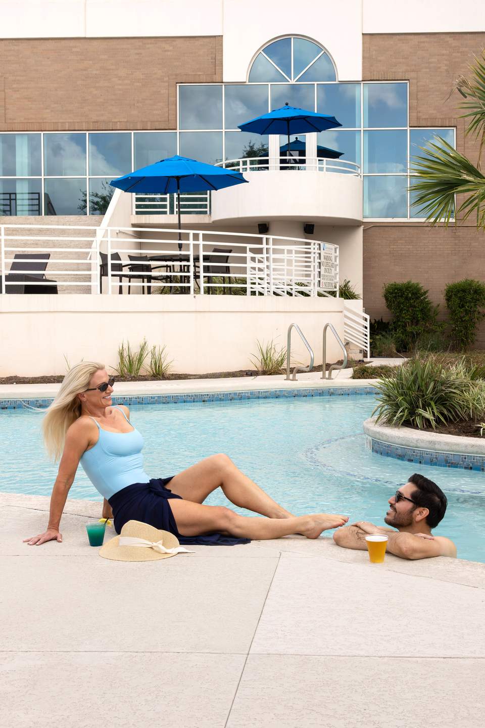 Woman and man relaxing poolside in front of a modern white building with blue umbrellas and glass balcony doors