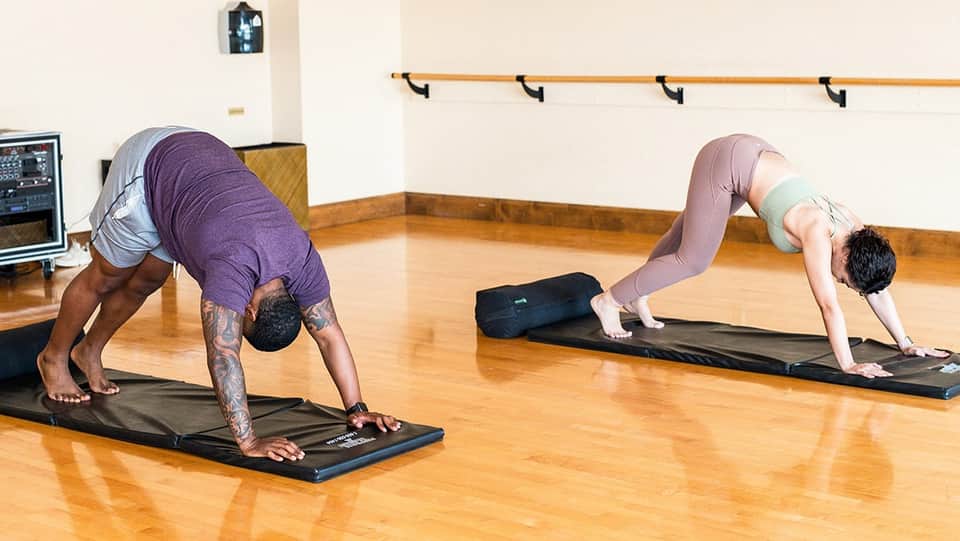 Yoga class with two instructors demonstrating downward dog pose on mats in a wooden studio with wall-mounted mirror