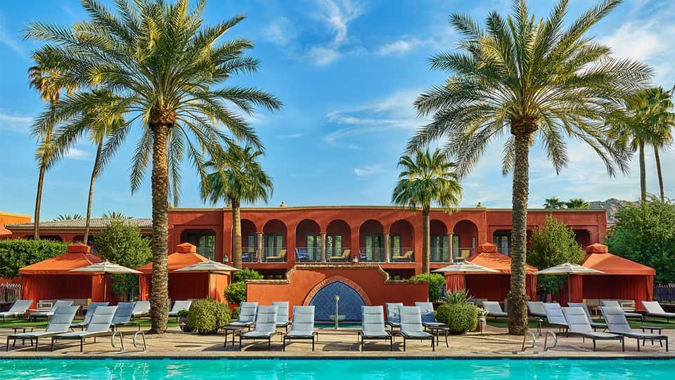 Resort pool with lounge chairs and orange umbrellas in front of red building with palm trees